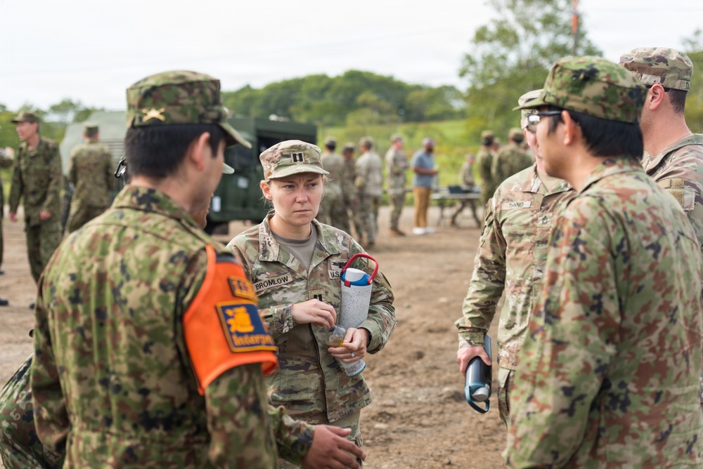 A group of military personnel in camouflage uniforms are gathered outdoors in what appears to be a joint military exercise or training event. A female U.S. Army Captain identified by her name tape as 'BROMLOW' is centrally positioned, holding a water bottle and appearing to brief or converse with personnel wearing different camouflage patterns. The presence of both U.S. Army OCP uniforms and what appear to be Japanese Ground Self-Defense Force uniforms suggests a bilateral or multinational military exercise.