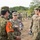 A group of military personnel in camouflage uniforms are gathered outdoors in what appears to be a joint military exercise or training event. A female U.S. Army Captain identified by her name tape as 'BROMLOW' is centrally positioned, holding a water bottle and appearing to brief or converse with personnel wearing different camouflage patterns. The presence of both U.S. Army OCP uniforms and what appear to be Japanese Ground Self-Defense Force uniforms suggests a bilateral or multinational military exercise.
