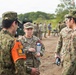 A group of military personnel in camouflage uniforms are gathered outdoors in what appears to be a joint military exercise or training event. A female U.S. Army Captain identified by her name tape as 'BROMLOW' is centrally positioned, holding a water bottle and appearing to brief or converse with personnel wearing different camouflage patterns. The presence of both U.S. Army OCP uniforms and what appear to be Japanese Ground Self-Defense Force uniforms suggests a bilateral or multinational military exercise.