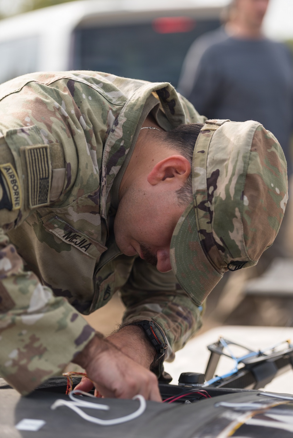 A U.S. Army Airborne soldier named Mejia is bent over, closely inspecting or working on what appears to be a drone or unmanned aerial system. The soldier is wearing Operational Camouflage Pattern (OCP) uniform with an Airborne tab, U.S. flag patch, and a name tape reading 'MEJIA'. The image captures focused technical work on electronic/wiring components of the equipment.