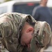 A U.S. Army Airborne soldier named Mejia is bent over, closely inspecting or working on what appears to be a drone or unmanned aerial system. The soldier is wearing Operational Camouflage Pattern (OCP) uniform with an Airborne tab, U.S. flag patch, and a name tape reading 'MEJIA'. The image captures focused technical work on electronic/wiring components of the equipment.