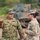 Two military personnel in different camouflage uniforms are engaged in conversation in an outdoor field environment. One soldier wearing Japanese Ground Self-Defense Force (JGSDF) pattern camouflage is gesturing while speaking to a second soldier in US Army Operational Camouflage Pattern (OCP) uniform. A tracked military vehicle is visible in the blurred background, suggesting a joint military exercise or training event.
