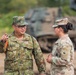 Two military personnel in different camouflage uniforms are engaged in conversation in an outdoor field environment. One soldier wearing Japanese Ground Self-Defense Force (JGSDF) pattern camouflage is gesturing while speaking to a second soldier in US Army Operational Camouflage Pattern (OCP) uniform. A tracked military vehicle is visible in the blurred background, suggesting a joint military exercise or training event.