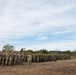 A large formation of military personnel in camouflage uniforms are assembled on an open dirt field, appearing to be at attention during a formal military ceremony or muster. The formation consists of multiple rows and columns of soldiers, with what appears to be a commanding officer or NCO standing in front addressing or inspecting the troops. The setting appears to be an outdoor military training area or base, with trees, military vehicles, and equipment visible in the background.