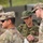The image shows a group of military personnel from at least two different armed forces engaged in a discussion or briefing outdoors. U.S. Army soldiers wearing OCP (Operational Camouflage Pattern) uniforms are interacting with what appears to be a Japan Ground Self-Defense Force (JGSDF) soldier in a distinctive Japanese camouflage uniform. The setting suggests a joint military exercise or coordination meeting.