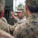A U.S. Army non-commissioned officer in OCP (Operational Camouflage Pattern) uniform is briefing or instructing a group of soldiers, including what appear to be Japanese Ground Self-Defense Force personnel based on their distinctive camouflage pattern. The NCO is gesturing with his right hand, suggesting he is actively explaining or demonstrating something near a piece of military equipment. The setting appears to be an outdoor military training or joint exercise environment.