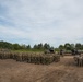 A large military formation is assembled on an open dirt field, with two distinct groups of soldiers visible — a larger group on the left and a smaller group on the right. Military vehicles, including what appear to be self-propelled artillery systems and support trucks, are positioned in the background. The setting appears to be a training area or exercise ground bordered by dense tree lines.