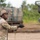 A U.S. Army soldier in full combat uniform is holding a wire or lanyard cord during what appears to be a field training exercise or live-fire operation. The soldier is wearing MultiCam (OCP) pattern uniform, a combat helmet with a mounted device, and tactical gloves. In the background, multiple large military vehicles — likely self-propelled artillery or rocket artillery systems — are visible in a wooded, sandy terrain environment.