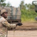 A U.S. Army soldier in full combat uniform is holding a wire or lanyard cord during what appears to be a field training exercise or live-fire operation. The soldier is wearing MultiCam (OCP) pattern uniform, a combat helmet with a mounted device, and tactical gloves. In the background, multiple large military vehicles — likely self-propelled artillery or rocket artillery systems — are visible in a wooded, sandy terrain environment.