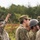A group of military personnel in camouflage uniforms are gathered outdoors in a wooded or field environment. One individual is holding a smartphone up high to capture a photo or video of something beyond the frame. The uniforms and cap patterns suggest these are Japanese Ground Self-Defense Force (JGSDF) members, with at least one individual wearing a different camouflage pattern.