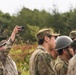 A group of military personnel in camouflage uniforms are gathered outdoors in a wooded or field environment. One individual is holding a smartphone up high to capture a photo or video of something beyond the frame. The uniforms and cap patterns suggest these are Japanese Ground Self-Defense Force (JGSDF) members, with at least one individual wearing a different camouflage pattern.