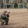 A person wearing military camouflage uniform and cap is crouched in a low squat position, photographing with a DSLR camera. They are wearing a blue armband/vest indicating a press or observer role. The setting appears to be a military training ground or exercise area with wooden barrier structures in the background.