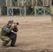 A person wearing military camouflage uniform and cap is crouched in a low squat position, photographing with a DSLR camera. They are wearing a blue armband/vest indicating a press or observer role. The setting appears to be a military training ground or exercise area with wooden barrier structures in the background.