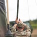 A soldier identified by the name tape 'TALBOT' on their patrol cap is working with a vertical pole or antenna mast, likely setting up or adjusting field communications equipment. The soldier is wearing US Army OCP (Operational Camouflage Pattern) uniform and a black wristwatch. The setting appears to be an outdoor military field environment with green military equipment visible nearby.