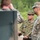 A Japan Ground Self-Defense Force (JGSDF) soldier, identified by his distinctive Japanese camouflage pattern and name tape reading 'KORU', appears to be briefing or instructing a group of U.S. Army soldiers wearing MultiCam/OCP uniforms. The setting appears to be an outdoor military training area with green vegetation in the background. This appears to be a joint military exercise or training event between Japanese and American forces.