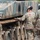 Two soldiers in MultiCam/OCP camouflage uniforms are inspecting or examining the track and running gear of a tracked military vehicle. One soldier, with hair in a bun and wearing a patrol cap, is touching the track links while the other observes. The vehicle appears to be a large armored tracked platform with visible road wheels, track assembly, and engine louvers.