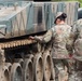 Two soldiers in MultiCam/OCP camouflage uniforms are inspecting or examining the track and running gear of a tracked military vehicle. One soldier, with hair in a bun and wearing a patrol cap, is touching the track links while the other observes. The vehicle appears to be a large armored tracked platform with visible road wheels, track assembly, and engine louvers.