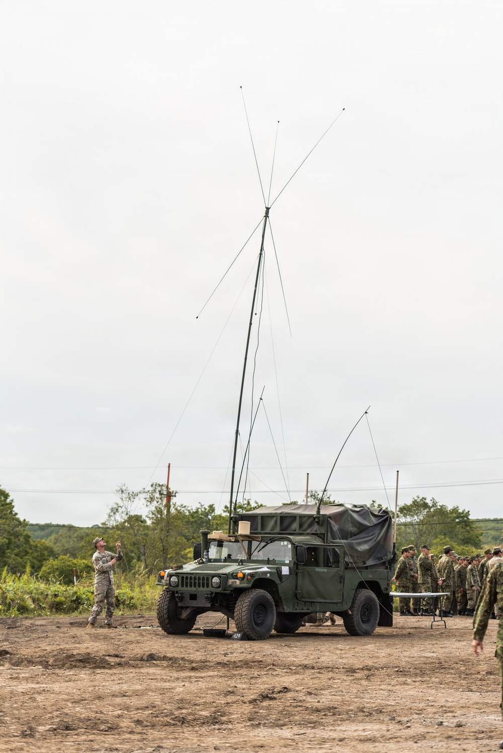 A military HMMWV (Humvee) is equipped with a tall erected antenna mast with multiple wire elements extending outward, consistent with a field communications setup. One soldier in camouflage uniform stands to the left observing or managing the antenna, while a group of uniformed personnel observes from the right background. The setting appears to be an open dirt field during an outdoor military training or demonstration exercise.