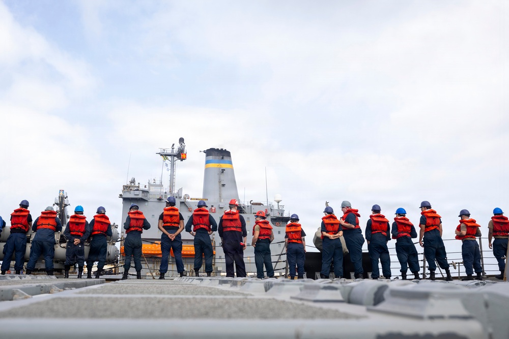 A group of naval personnel wearing orange life vests and blue/red hard hats are lined up along the deck railing of a military vessel, facing a larger gray ship alongside them. The personnel appear to be in formation during what is likely a replenishment-at-sea (RAS) or ship-to-ship operation. The larger vessel displays a distinctive funnel with blue and yellow bands, characteristic of a naval auxiliary or replenishment ship.