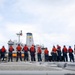 A group of naval personnel wearing orange life vests and blue/red hard hats are lined up along the deck railing of a military vessel, facing a larger gray ship alongside them. The personnel appear to be in formation during what is likely a replenishment-at-sea (RAS) or ship-to-ship operation. The larger vessel displays a distinctive funnel with blue and yellow bands, characteristic of a naval auxiliary or replenishment ship.