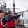 Delbert D. Black Conducts Replenishment-at-Sea with USNS John Lenthall Two naval personnel aboard a warship are conducting a line-throwing operation at sea, with one sailor firing a line-throwing gun (rifle-type launcher) toward another vessel. Both are wearing standard naval safety gear including hard hats, life vests, and eye protection. A second warship is visible in the background, suggesting a replenishment-at-sea (RAS) or ship-to-ship transfer evolution.