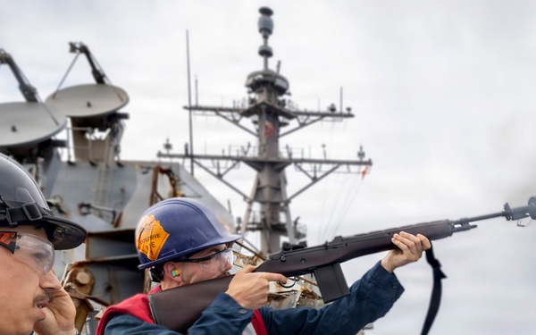Two naval personnel aboard a warship are conducting a line-throwing operation at sea, with one sailor firing a line-throwing gun (rifle-type launcher) toward another vessel. Both are wearing standard naval safety gear including hard hats, life vests, and eye protection. A second warship is visible in the background, suggesting a replenishment-at-sea (RAS) or ship-to-ship transfer evolution.