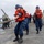 Delbert D. Black Conducts Replenishment-at-Sea with USNS John Lenthall A group of U.S. Navy sailors are performing line-handling operations on the deck of a naval vessel at sea. The foreground sailor, wearing a dark uniform with a U.S. flag patch, is pulling a thick rope while wearing an orange life vest and blue hard hat. Multiple sailors in identical safety gear are lined up behind her in a coordinated team effort.