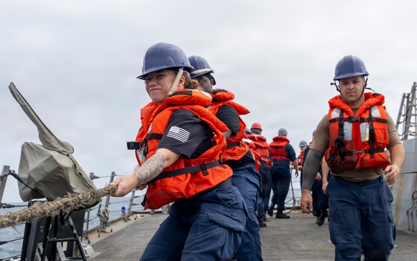 A group of U.S. Navy sailors are performing line-handling operations on the deck of a naval vessel at sea. The foreground sailor, wearing a dark uniform with a U.S. flag patch, is pulling a thick rope while wearing an orange life vest and blue hard hat. Multiple sailors in identical safety gear are lined up behind her in a coordinated team effort.