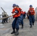 A group of U.S. Navy sailors are performing line-handling operations on the deck of a naval vessel at sea. The foreground sailor, wearing a dark uniform with a U.S. flag patch, is pulling a thick rope while wearing an orange life vest and blue hard hat. Multiple sailors in identical safety gear are lined up behind her in a coordinated team effort.