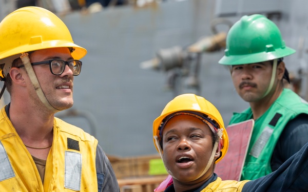 Three workers are visible on what appears to be a naval vessel or industrial maritime setting. Two workers in yellow hard hats and yellow safety vests are in the foreground, with one gesturing and appearing to give directions or instructions. A third worker in a green hard hat and green vest stands in the background.