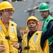 Three workers are visible on what appears to be a naval vessel or industrial maritime setting. Two workers in yellow hard hats and yellow safety vests are in the foreground, with one gesturing and appearing to give directions or instructions. A third worker in a green hard hat and green vest stands in the background.