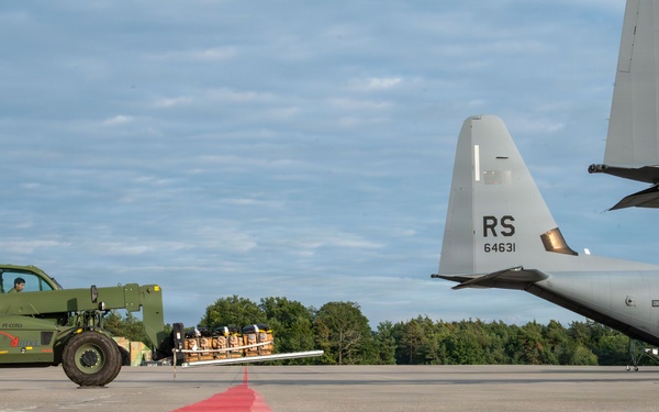 A military airfield ramp scene showing U.S. Air Force C-130 Hercules aircraft with tail codes 'RS 85683' and 'RS 64631', indicating assignment to Ramstein Air Base, Germany. A military green rough terrain forklift (telehandler) is transporting a pallet of cargo across the tarmac toward the aircraft. The low-angle ground-level shot emphasizes a red painted boundary line on the concrete ramp surface.