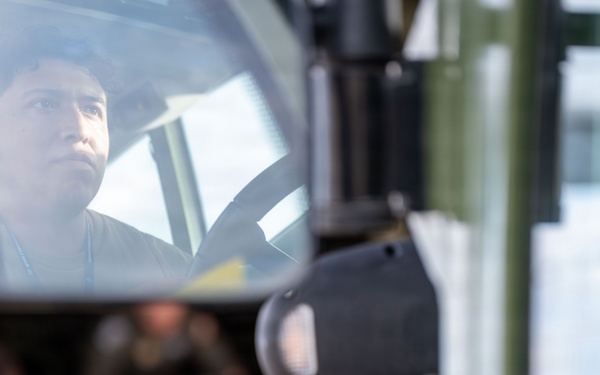 A close-up photograph captures a person's reflection in the side mirror of a military-style green vehicle. The composition uses selective focus, with the mirror reflection sharp and the background blurred, showing additional personnel and cargo in the background. The vehicle appears to be a large military or tactical truck.