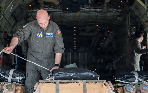 A U.S. Air Force loadmaster in olive drab flight suit is working at the open rear cargo ramp of a military transport aircraft, likely a C-130 Hercules. He is handling a static line or strap connected to cargo bundles loaded on the ramp. The cargo consists of airdrop bundles secured with straps, honeycomb cardboard cushioning, and what appear to be water containers or fuel bladders labeled 'DEMOC'.
