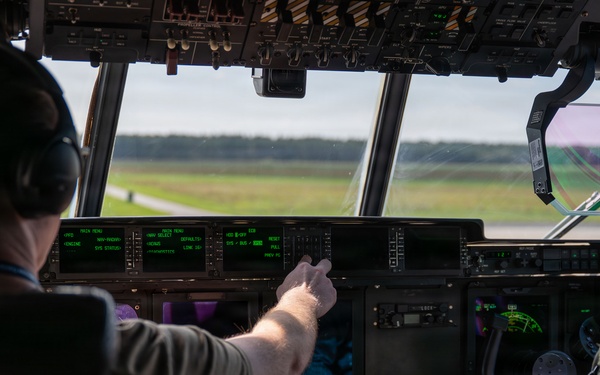 Two military aircrew members are seated in the cockpit of a large military transport aircraft, likely a C-17 Globemaster III or similar aircraft, on a runway or taxiway. The left-seat pilot is pointing at the green-lit instrument panel displays while the right-seat crew member, wearing a flight suit with a visible U.S. flag patch, observes. The aircraft appears to be on the ground with a runway and tree line visible through the windscreen.