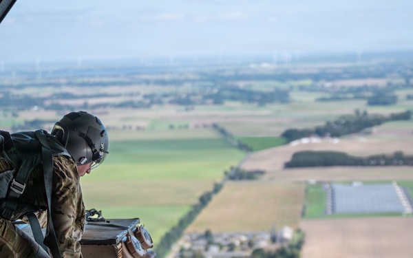 A military loadmaster or aircrew member is positioned at the open rear cargo ramp of a military transport aircraft, leaning over a secured cargo pallet at altitude. The aircraft is flying over a flat, agricultural landscape with visible fields, tree lines, roads, and what appears to be a solar panel installation. The image captures an aerial cargo operation, likely a airdrop preparation or cargo inspection.