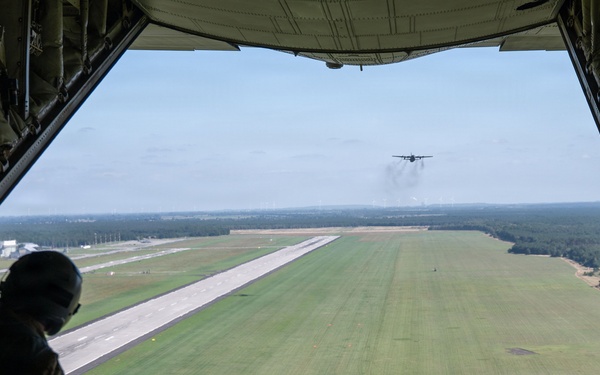 A military loadmaster or crew member stands at the open rear cargo ramp of a large military transport aircraft, likely a C-130 Hercules, observing an airdrop operation over an airfield. A parachute-deployed bundle or container can be seen mid-air above the runway below, while a second military transport aircraft is visible in the distance approaching or departing the runway. The image is taken from inside the aircraft looking outward through the open ramp.