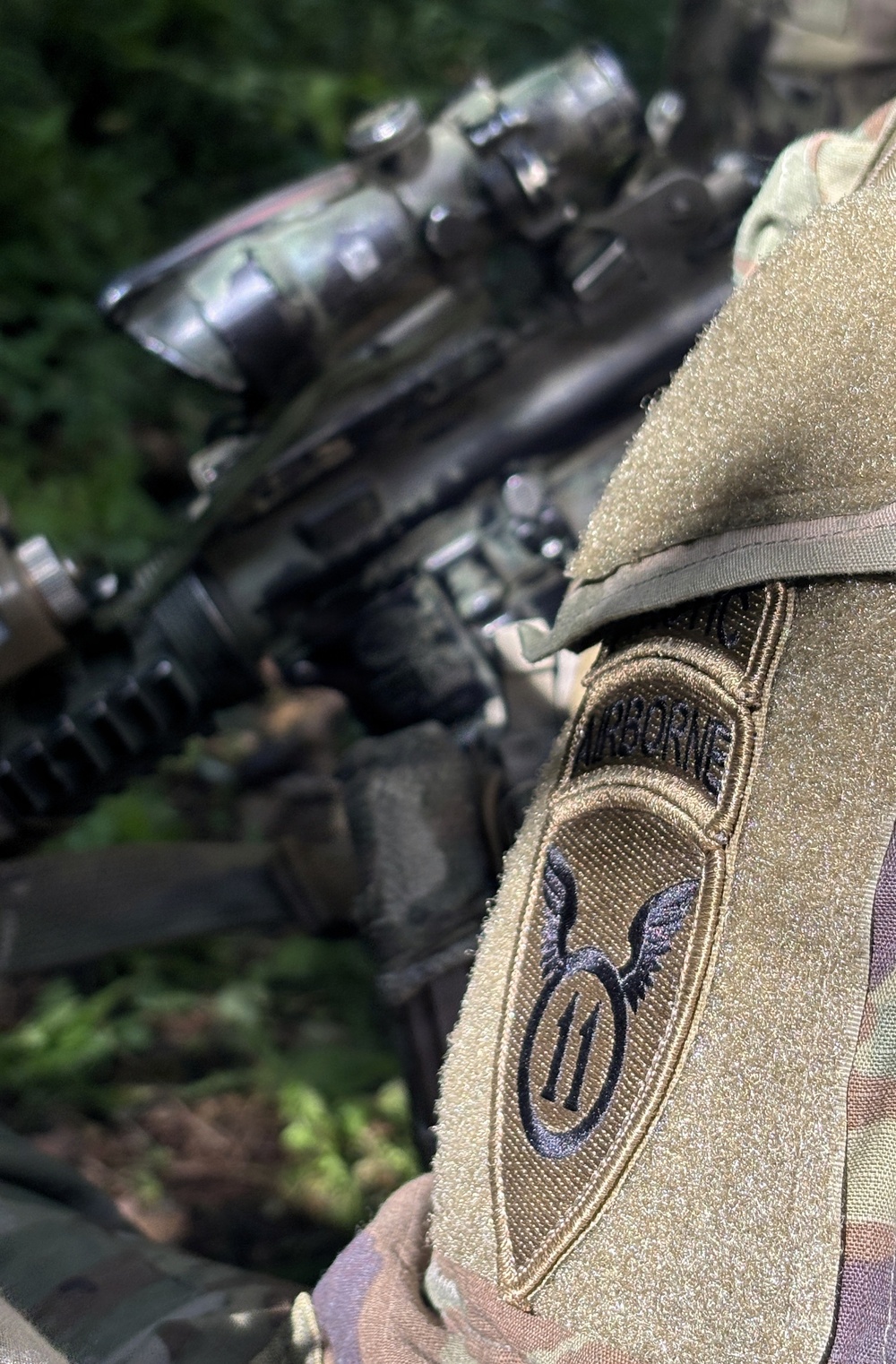 Close-up photograph of a U.S. Army soldier's shoulder sleeve insignia showing the 11th Airborne Division patch on a MultiCam/OCP camouflage uniform. The patch features the division's distinctive winged numeral '11' design with an 'AIRBORNE' tab above it. A rifle with optics is visible in the blurred background.