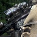 Close-up photograph of a U.S. Army soldier's shoulder sleeve insignia showing the 11th Airborne Division patch on a MultiCam/OCP camouflage uniform. The patch features the division's distinctive winged numeral '11' design with an 'AIRBORNE' tab above it. A rifle with optics is visible in the blurred background.