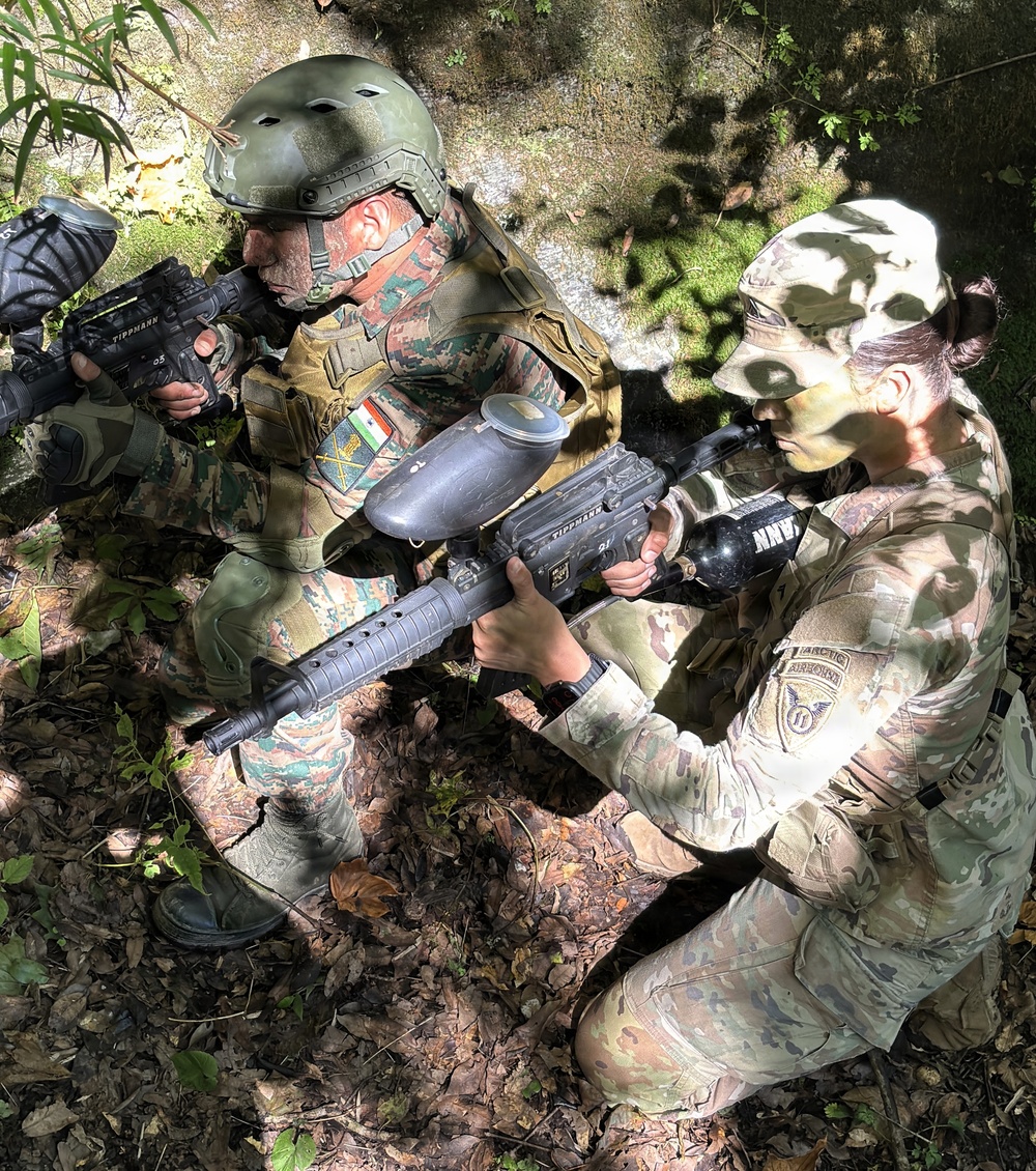 Two individuals in military-style camouflage uniforms are crouching in a wooded outdoor setting, aiming Tippmann paintball markers. The person on the left wears a tactical helmet and a patch resembling an Indian flag, while the person on the right wears a cap with what appears to be an Airborne unit patch. This appears to be a paintball or airsoft recreational activity staged in a realistic military simulation format.