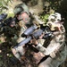 Two individuals in military-style camouflage uniforms are crouching in a wooded outdoor setting, aiming Tippmann paintball markers. The person on the left wears a tactical helmet and a patch resembling an Indian flag, while the person on the right wears a cap with what appears to be an Airborne unit patch. This appears to be a paintball or airsoft recreational activity staged in a realistic military simulation format.