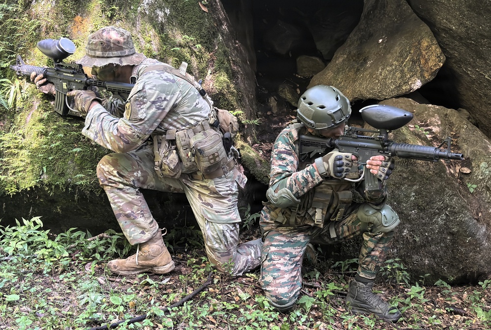 Two individuals in full military-style camouflage gear are positioned behind large moss-covered rocks in a forested outdoor environment. Both are holding paintball markers (guns) with hopper feed systems visible on top, indicating this is a paintball or milsim (military simulation) activity. They appear to be taking tactical cover positions as part of a scenario-based game.