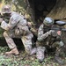 Two individuals in full military-style camouflage gear are positioned behind large moss-covered rocks in a forested outdoor environment. Both are holding paintball markers (guns) with hopper feed systems visible on top, indicating this is a paintball or milsim (military simulation) activity. They appear to be taking tactical cover positions as part of a scenario-based game.