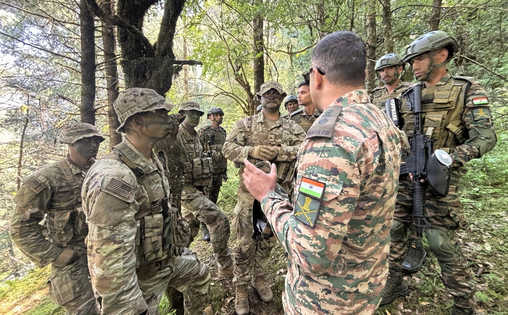 A group of military personnel from at least two nations are gathered in a forested area, appearing to be engaged in a briefing or debriefing session. Soldiers wearing US Army MultiCam uniforms and Indian Army camouflage uniforms are visible, suggesting a joint military exercise or training operation. An Indian Army soldier facing away from the camera appears to be addressing the assembled group.