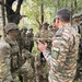 A group of military personnel from at least two nations are gathered in a forested area, appearing to be engaged in a briefing or debriefing session. Soldiers wearing US Army MultiCam uniforms and Indian Army camouflage uniforms are visible, suggesting a joint military exercise or training operation. An Indian Army soldier facing away from the camera appears to be addressing the assembled group.