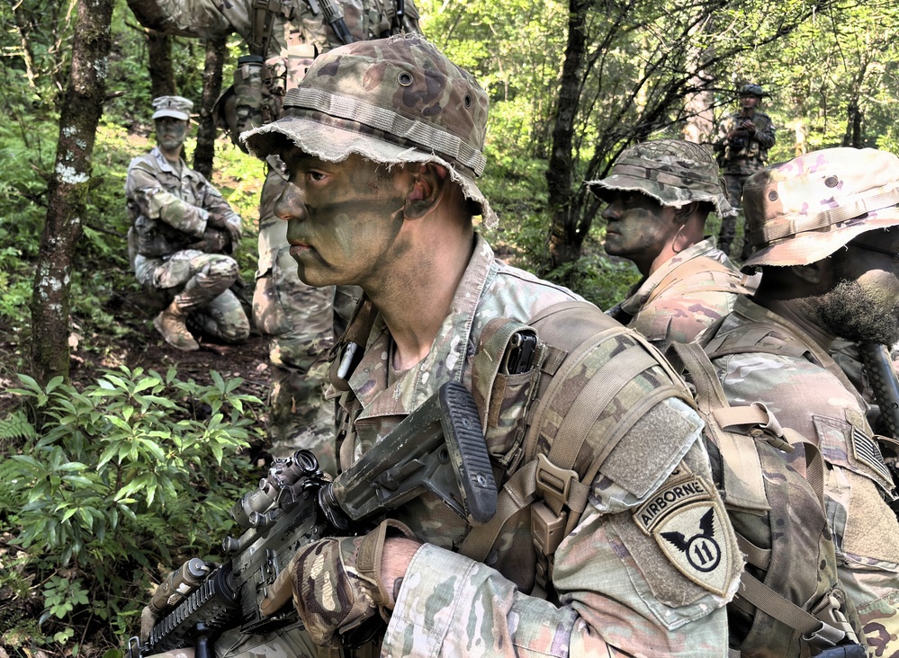 The image shows a group of U.S. Army soldiers in a forested environment wearing MultiCam (OCP) uniforms with camouflage face paint and boonie hats. The foreground soldier displays an '11th Airborne Division' patch on their left shoulder and is carrying an M4-style carbine. The scene appears to be a field training exercise or patrol operation in a wooded, hilly terrain.