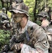 The image shows a group of U.S. Army soldiers in a forested environment wearing MultiCam (OCP) uniforms with camouflage face paint and boonie hats. The foreground soldier displays an '11th Airborne Division' patch on their left shoulder and is carrying an M4-style carbine. The scene appears to be a field training exercise or patrol operation in a wooded, hilly terrain.