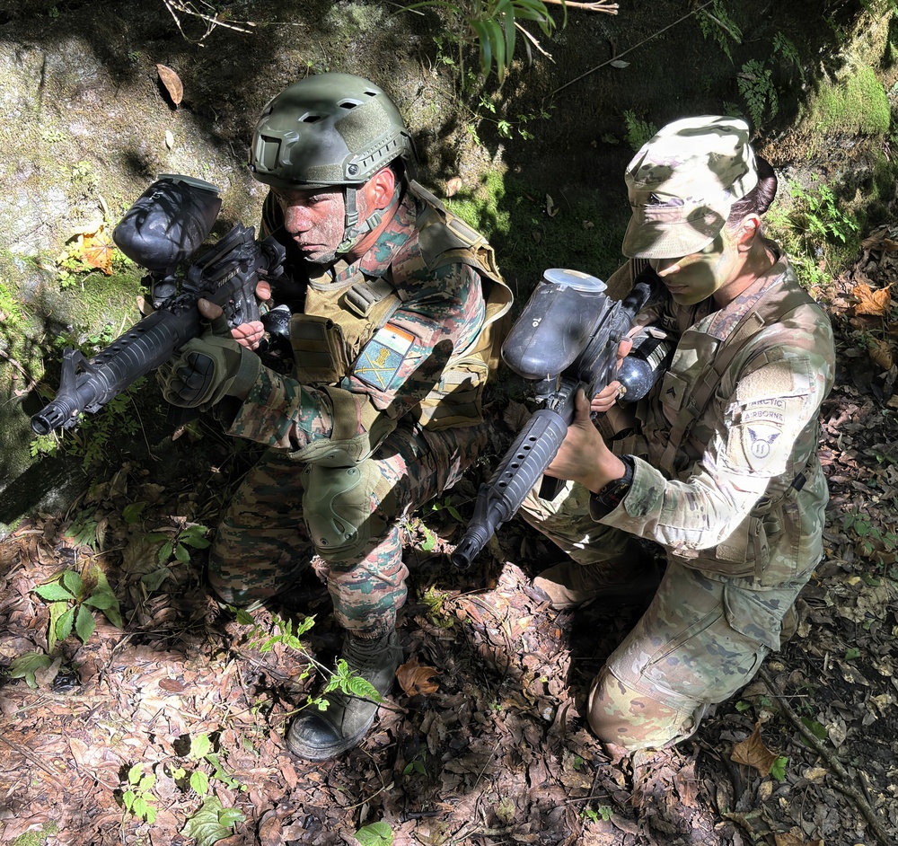 Two individuals in military-style camouflage uniforms are crouching in a wooded outdoor setting, holding paintball or airsoft markers with hopper attachments. Both have face paint applied and are wearing tactical gear including vests and knee pads. The scene appears to be a recreational airsoft or paintball skirmish game.