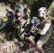 Two individuals in military-style camouflage uniforms are crouching in a wooded outdoor setting, holding paintball or airsoft markers with hopper attachments. Both have face paint applied and are wearing tactical gear including vests and knee pads. The scene appears to be a recreational airsoft or paintball skirmish game.