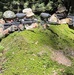 Three individuals in military-style camouflage uniforms are positioned behind a large moss-covered rock in a forested environment, aiming paintball guns forward. They appear to be engaged in a paintball or airsoft training exercise, as evidenced by the visible hopper/loader attachments on the weapons. The setting is a dense woodland area with pine trees and dappled sunlight.